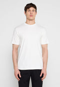 Mann steht mit dem Gesicht nach vorne, trägt ein schlichtes weißes T-Shirt mit kurzen Ärmeln und schwarze Hosen vor einem einfarbigen hellen Hintergrund.