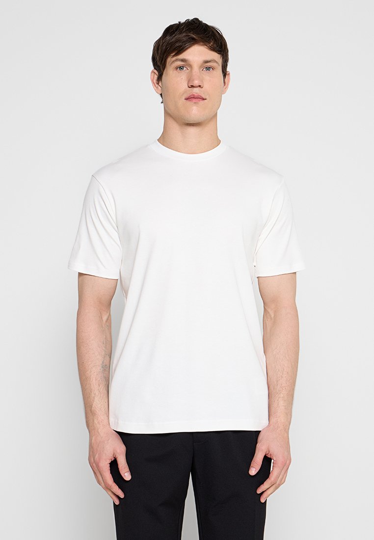 Mann steht mit dem Gesicht nach vorne, trägt ein schlichtes weißes T-Shirt mit kurzen Ärmeln und schwarze Hosen vor einem einfarbigen hellen Hintergrund.