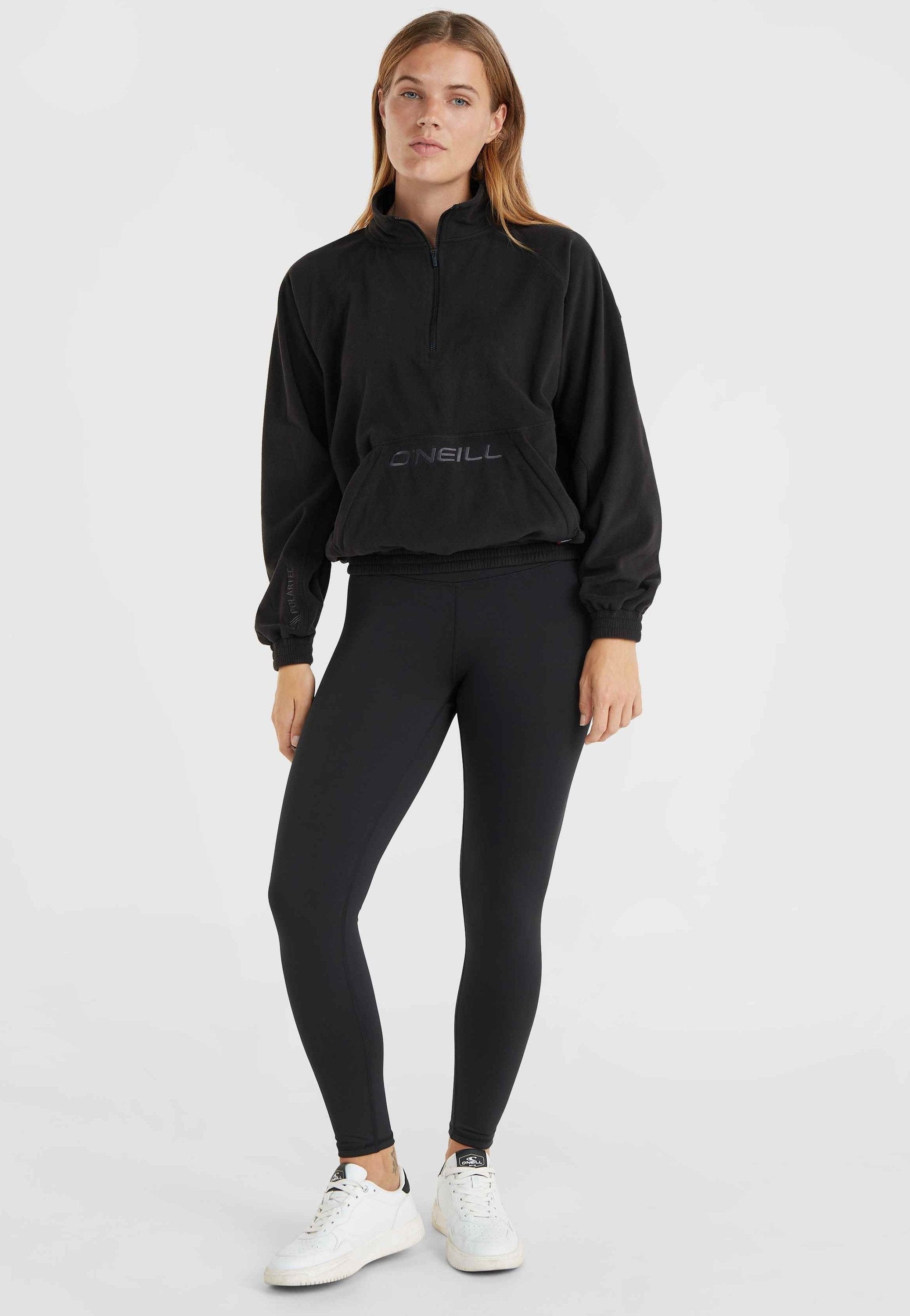Schwarzer Fleece-Pullover mit Viertelreißverschluss und Logo auf der Vorderseite, kombiniert mit schwarzen Leggings und weißen Sneakern. Elastische Bündchen und Taillenbund.