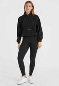 Schwarzer Fleece-Pullover mit Viertelreißverschluss und Logo auf der Vorderseite, kombiniert mit schwarzen Leggings und weißen Sneakern. Elastische Bündchen und Taillenbund.