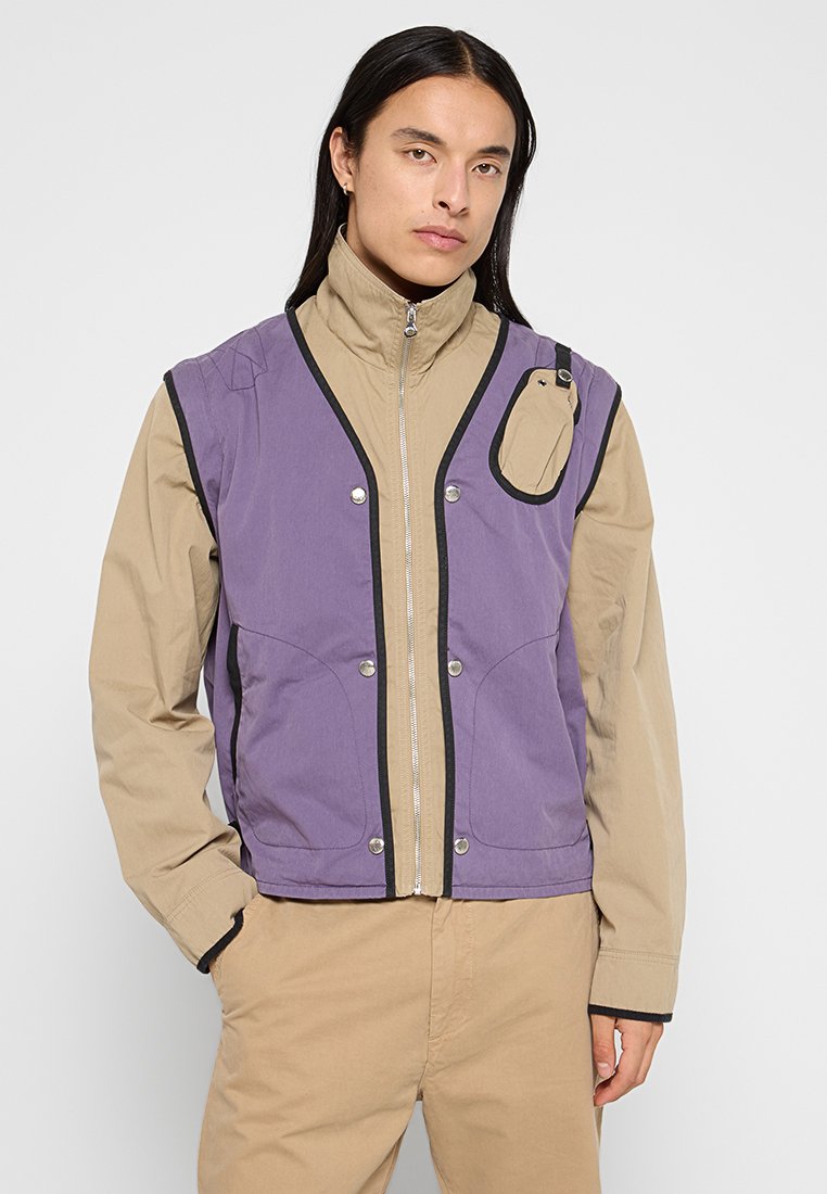 Lila- und beige Zip-Jacke mit hohem Kragen, kontrastierenden schwarzen Akzenten, zwei seitlichen Taschen und einem kleinen Detail auf der Brusttasche.