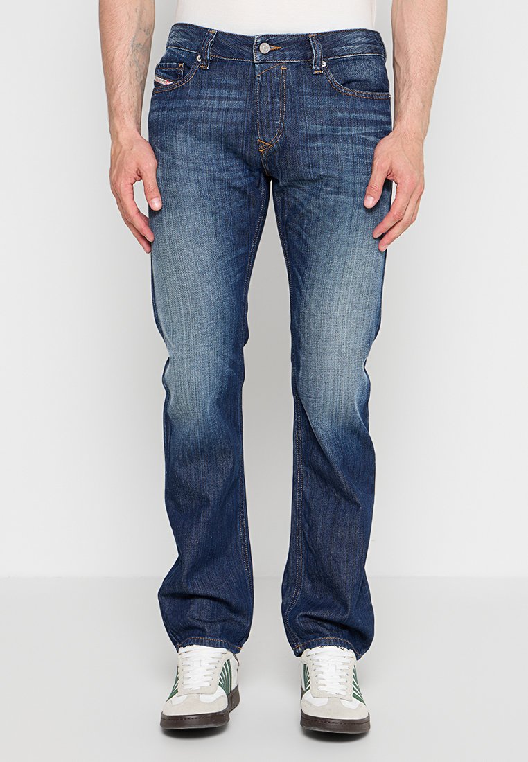 Mann trägt ausgebleichte blaue Jeans mit geradem Bein und weiße Sneaker, steht vor einem schlichten hellen Hintergrund.
