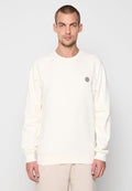 Mann, der einen schlichten, cremefarbenen Langarm-Sweatshirt trägt, mit einem kleinen runden Logo auf der Brust, vor einem weißen Hintergrund stehend.