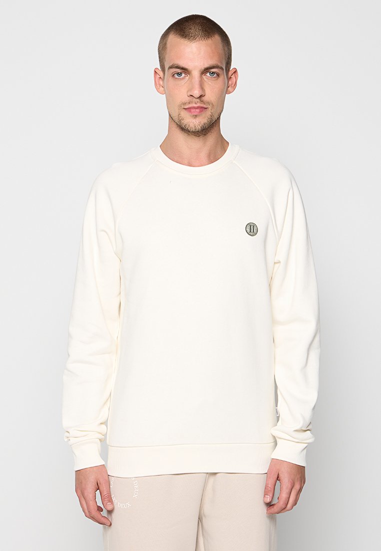Mann, der einen schlichten, cremefarbenen Langarm-Sweatshirt trägt, mit einem kleinen runden Logo auf der Brust, vor einem weißen Hintergrund stehend.
