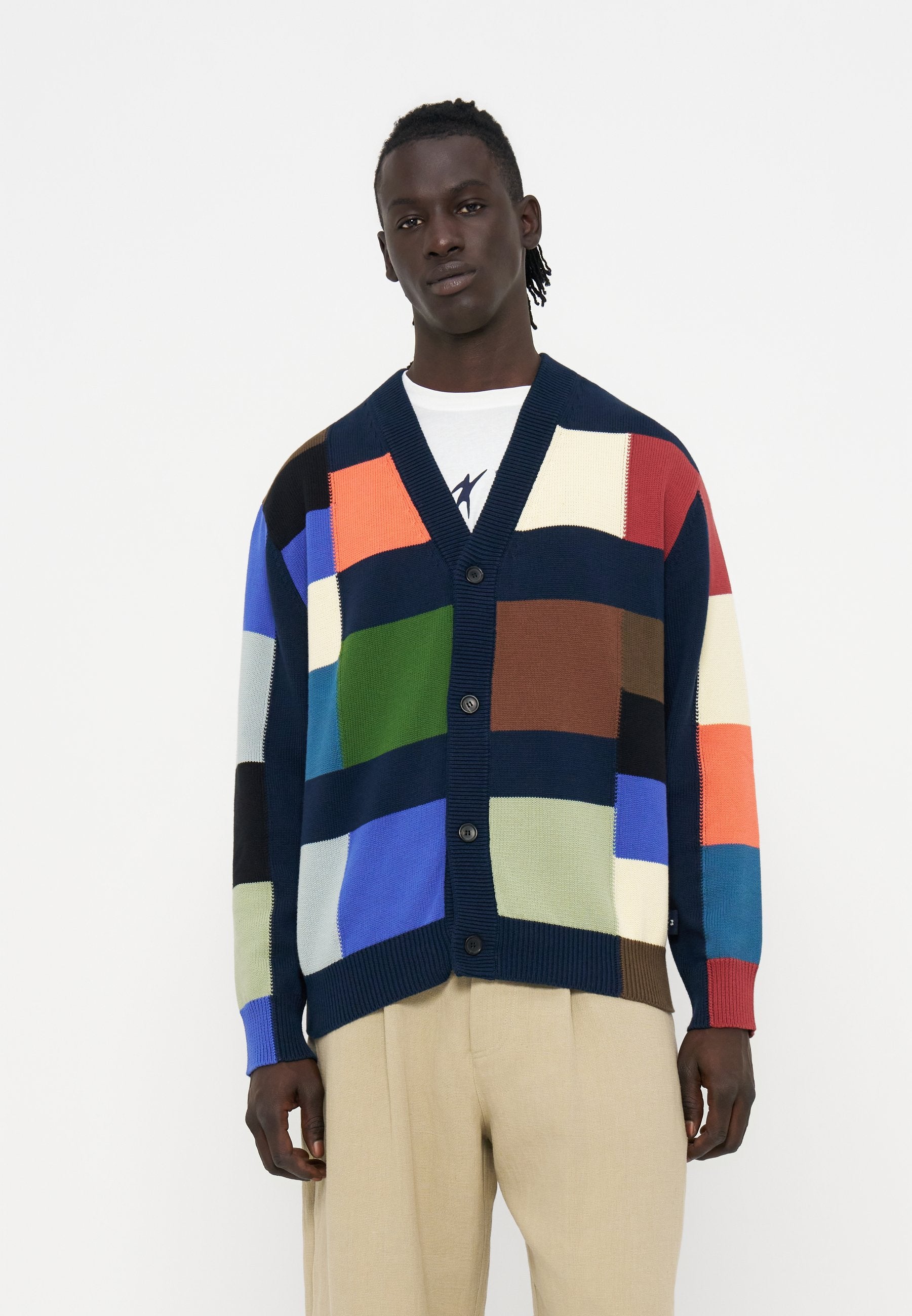 Mann trägt eine mehrfarbige Patchwork-Strickjacke mit Knöpfen über einem weißen Hemd und beigen Hosen vor einem einfarbigen Hintergrund.