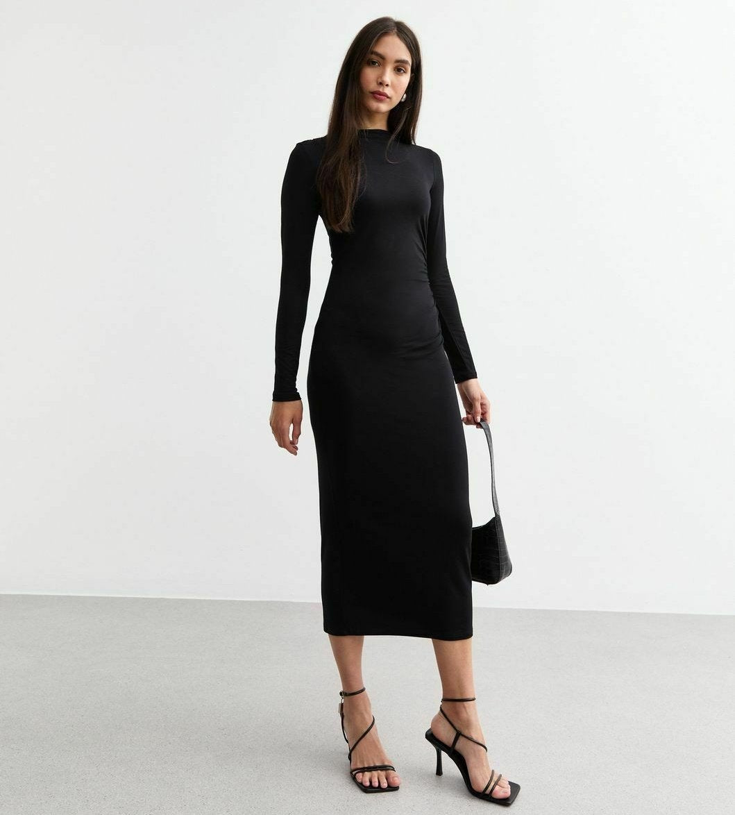 Schwarzes, langärmeliges, figurbetontes Kleid aus dehnbarem Stoff, knielang mit Rundhalsausschnitt. Das Modell hält eine kleine schwarze Handtasche und trägt Riemchensandalen.