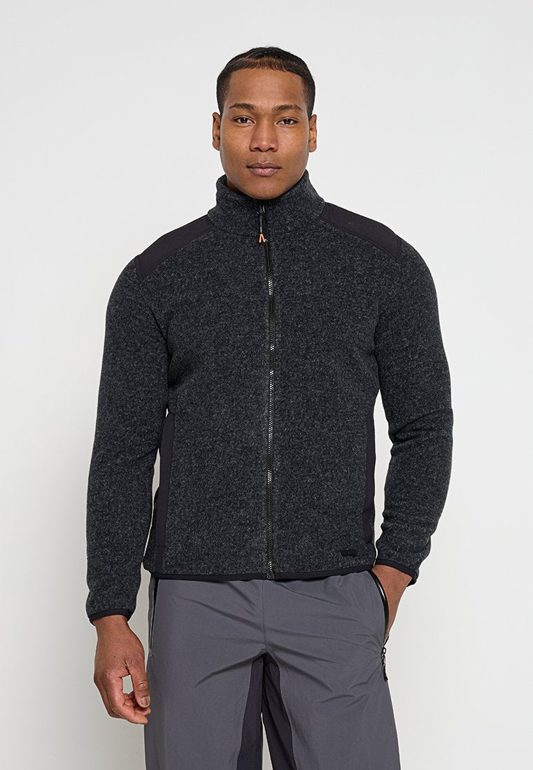 Mann trägt eine dunkelgraue Fleecejacke mit Reißverschluss und graue Hose, steht mit einer Hand in der Tasche vor einem schlichten weißen Hintergrund.
