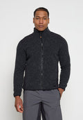 Mann trägt eine dunkelgraue Fleecejacke mit Reißverschluss und graue Hose, steht mit einer Hand in der Tasche vor einem schlichten weißen Hintergrund.