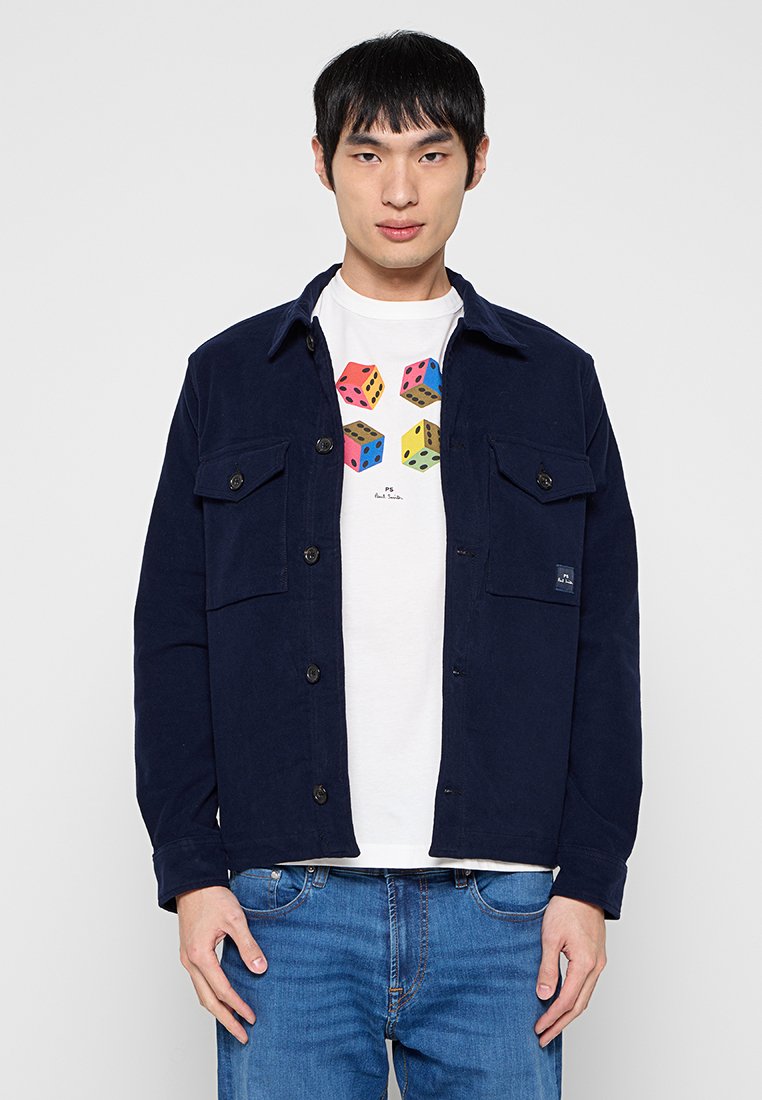 Marineblaue Cordjacke aus Cord mit zwei Brusttaschen, Knopfleiste und lockerem Schnitt, getragen über einem weißen T-Shirt mit Grafik und blauer Jeans.