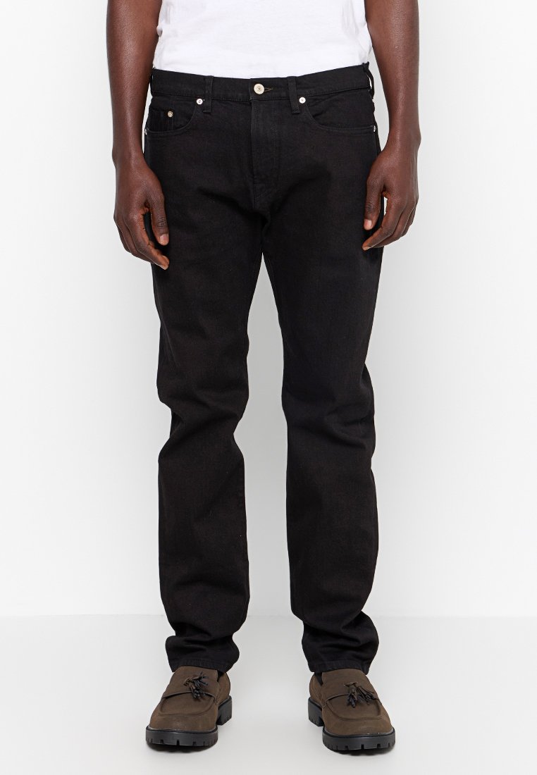 Schwarze Jeans aus Denim mit geradem Schnitt, fünf Taschen-Design, vorderem Knopfverschluss und silbernen Details. Getragen mit dunkelbraunen Schuhen.