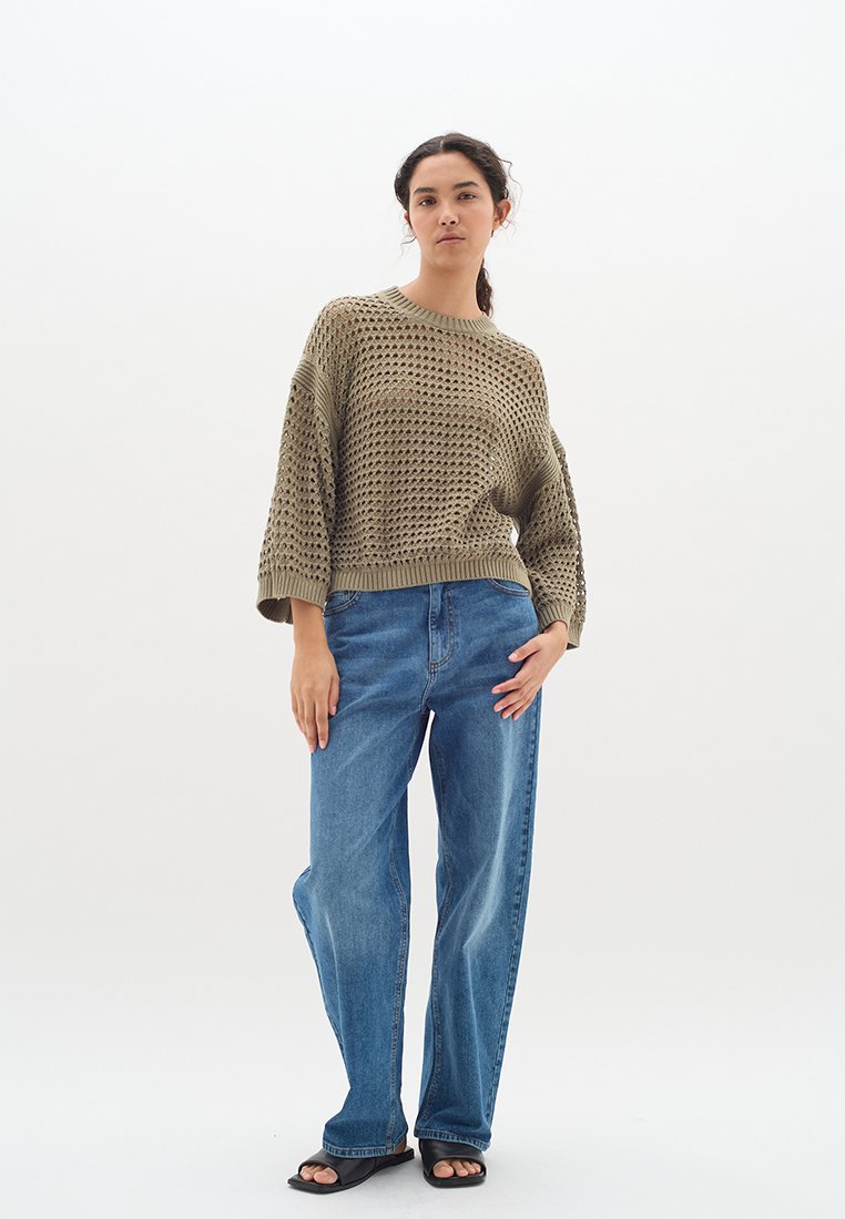 Olivgrüner Strickpullover mit einem lockeren, strukturierten Design, kombiniert mit hellblauen weit geschnittenen Jeans und schwarzen Sandalen.