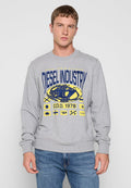 Junger Mann in grauem Sweatshirt mit "Diesel Industry" und grafischen Logos in Blau und Gelb, kombiniert mit blauen Jeans vor einem schlichten hellen Hintergrund.