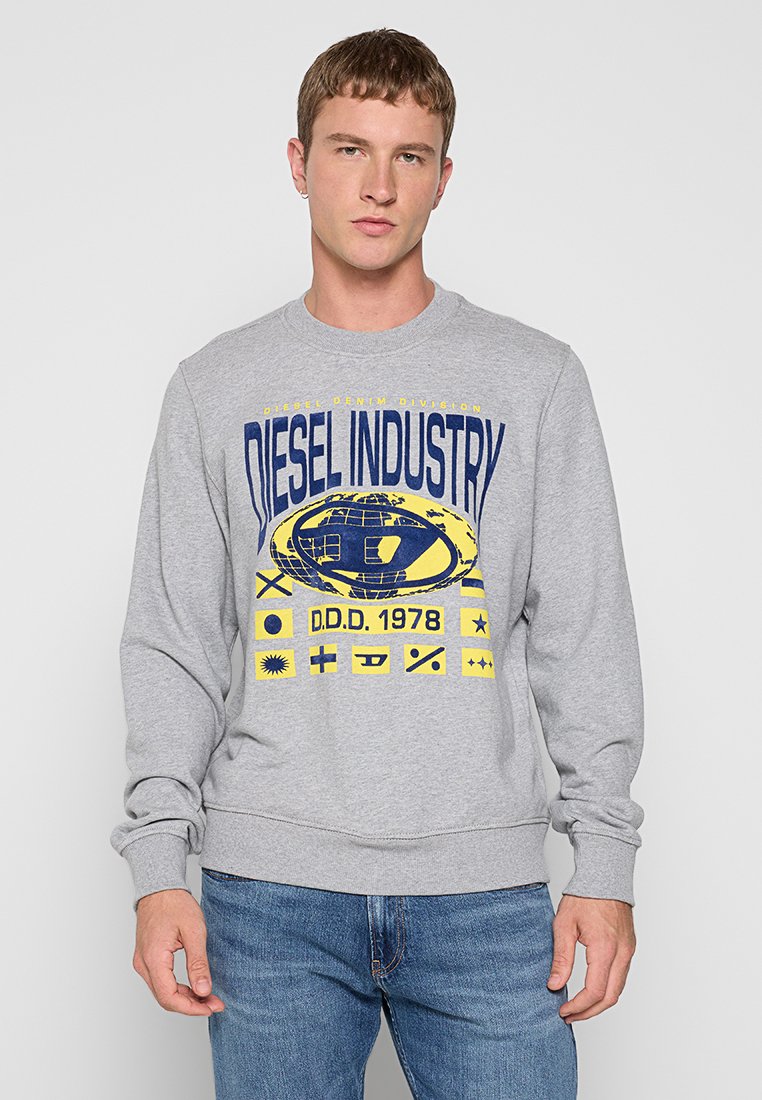 Junger Mann in grauem Sweatshirt mit "Diesel Industry" und grafischen Logos in Blau und Gelb, kombiniert mit blauen Jeans vor einem schlichten hellen Hintergrund.