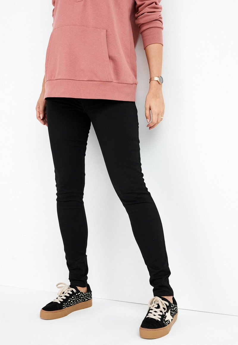 Schwarze Slim-Fit-Jeans, pinke Kapuzenjacke mit frontaler Tasche, schwarze Sneaker mit strukturiertem Muster und beigen Sohlen, die eine figurbetonte Silhouette präsentieren.