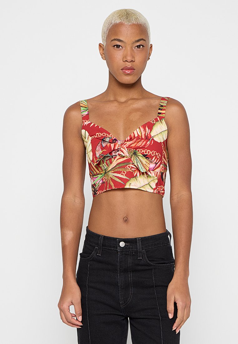 Person mit kurzen blonden Haaren trägt ein rotes Croptop mit tropischen Blatt- und Kettenmustern, kombiniert mit hoch taillierten schwarzen Jeans.