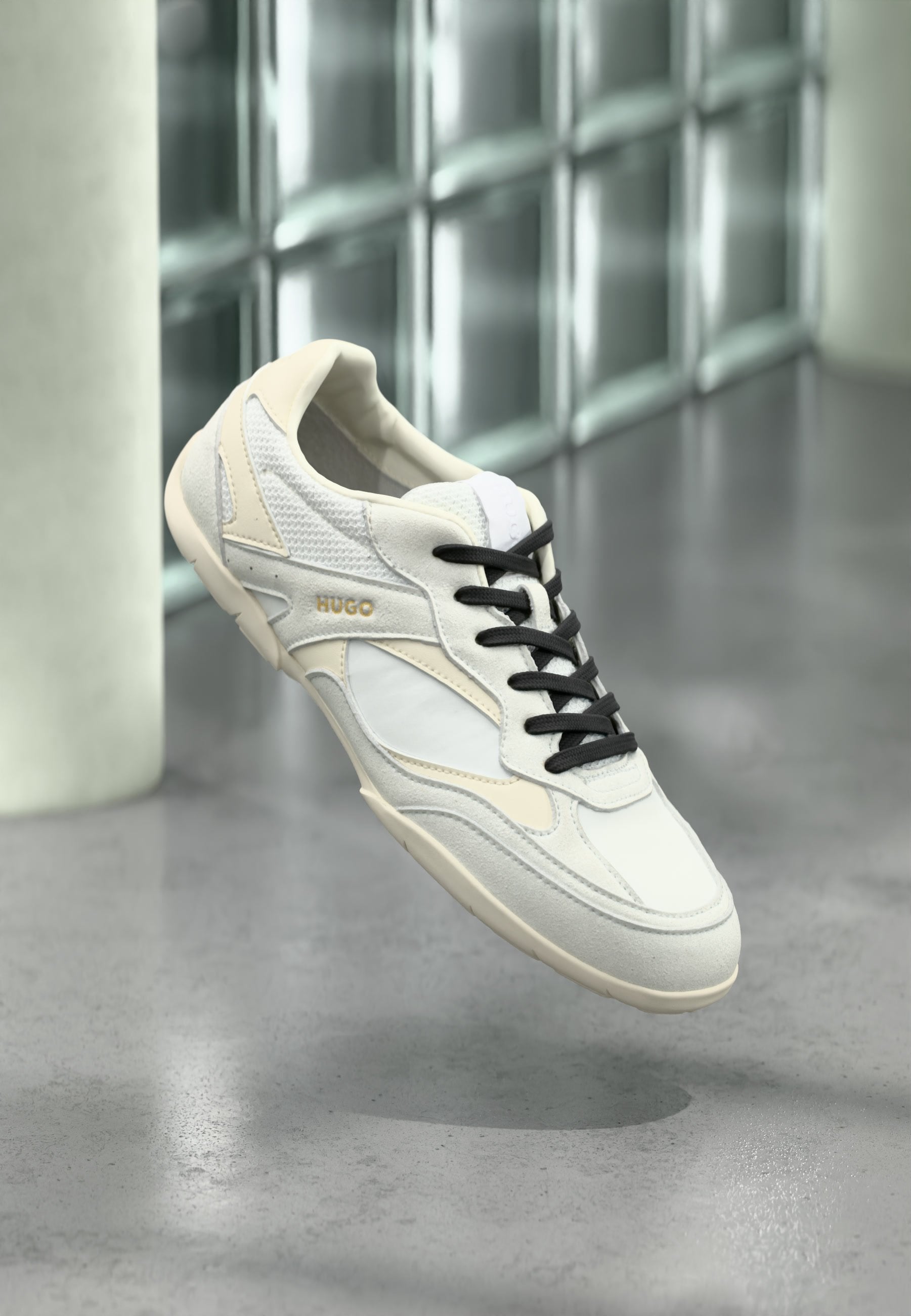Weißer und beiger Sneaker mit schwarzen Schnürsenkeln und "HUGO"-Markenlogo, der über einem polierten grauen Boden mit Hintergrund aus Glassteinwand schwebt.