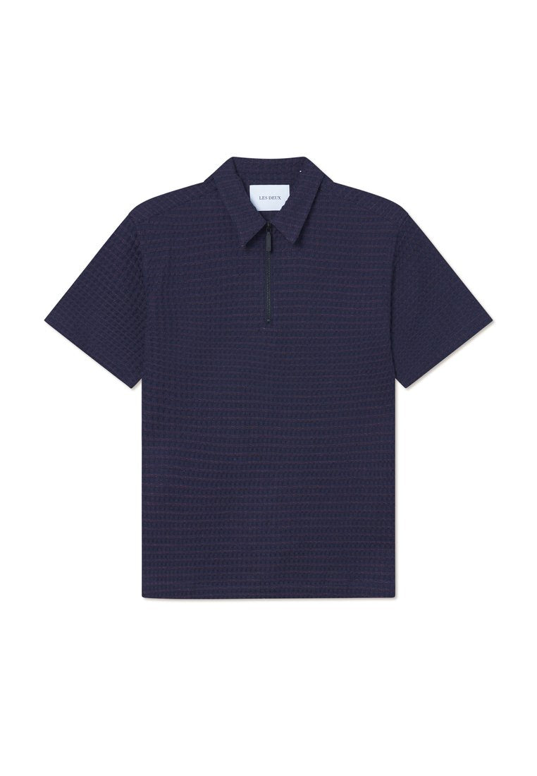 Marineblaues Polohemd mit kurzen Ärmeln und einem halben Reißverschluss am Kragen, strukturierter Stoff und subtil gemustertes geometrisches Design. Auf dem Kragen befindet sich ein Etikett mit der Aufschrift "LES DEUX."