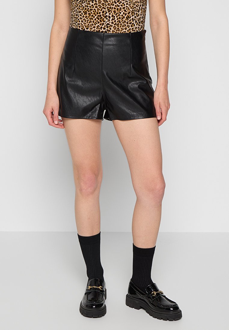 Schwarze Leder-Shorts mit glatter Textur, hoher Taille und tailliertem Schnitt. Kombiniert mit schwarzen Loafers und knielangen schwarzen Socken.