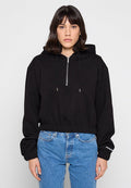 Frau trägt ein schwarzes, kurzes Hoodie mit Reißverschluss vorne und blaue Jeans, steht vor einem hellgrauen Hintergrund.