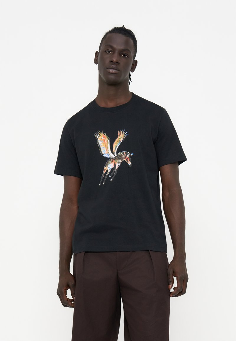 Ein Mann trägt ein schwarzes T-Shirt mit einem bunten, geflügelten Zebra-Motiv und dunkelbraunen Hosen und steht vor einem schlichten weißen Hintergrund.