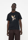 Ein Mann trägt ein schwarzes T-Shirt mit einem bunten, geflügelten Zebra-Motiv und dunkelbraunen Hosen und steht vor einem schlichten weißen Hintergrund.