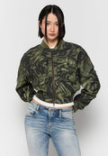 Junge Frau mit glattem schwarzen Haar trägt eine grüne Camouflage-Bomberjacke mit mehreren Vorderaschen und hellblauen verwaschenen Jeans.