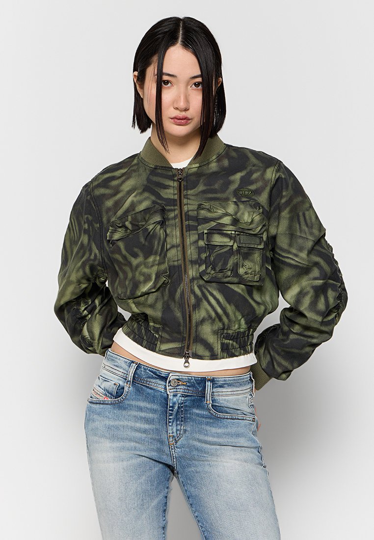 Junge Frau mit glattem schwarzen Haar trägt eine grüne Camouflage-Bomberjacke mit mehreren Vorderaschen und hellblauen verwaschenen Jeans.