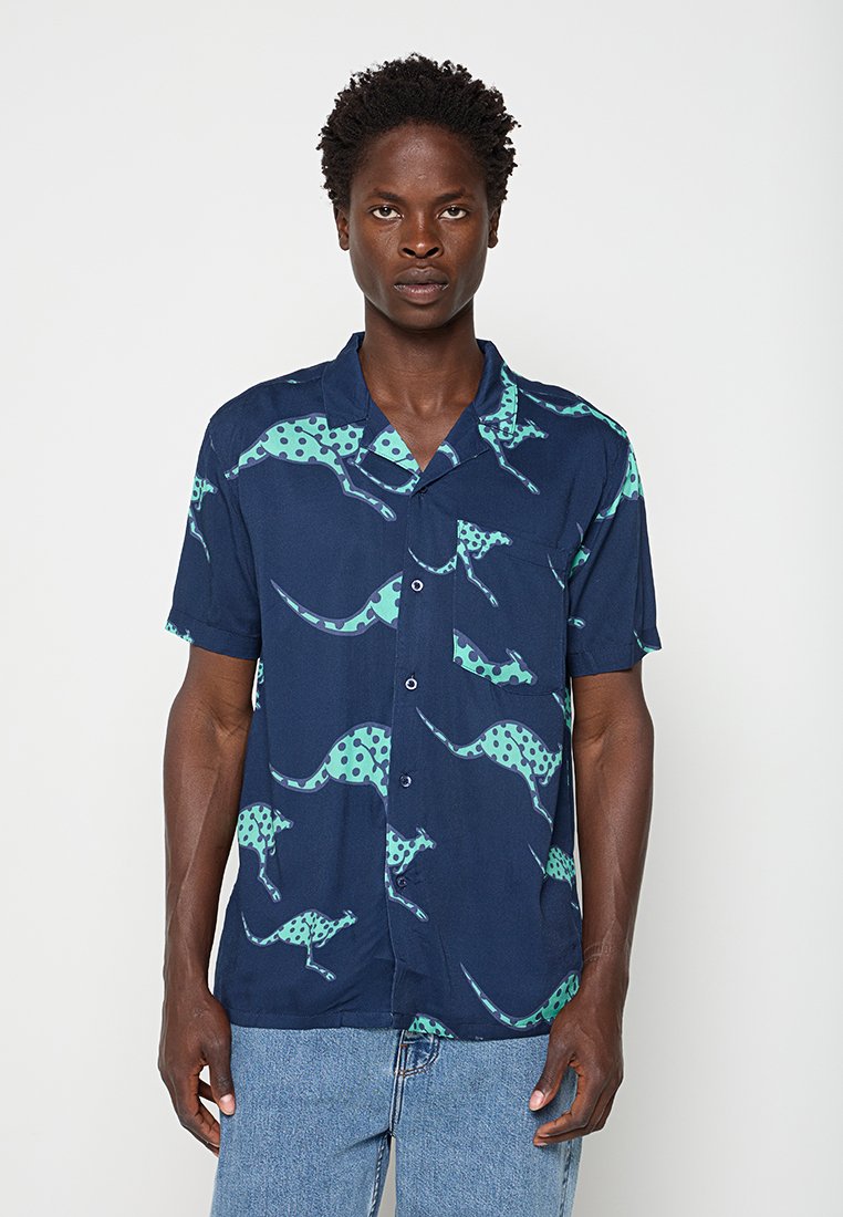 Mann trägt ein kurzärmeliges, marineblaues Hemd mit türkisfarbenen, gepunkteten Känguru-Prints und hellblauen Jeans.