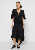 Schwarzes Kleid mit lockeren Ärmeln, V-Ausschnitt und gerafftem Bund. Mit einem ausgestellten Rock mit einem hoch-niedrig geschnittenen Saum und strukturierter Stoff. Schwarze Schnürschuhe.