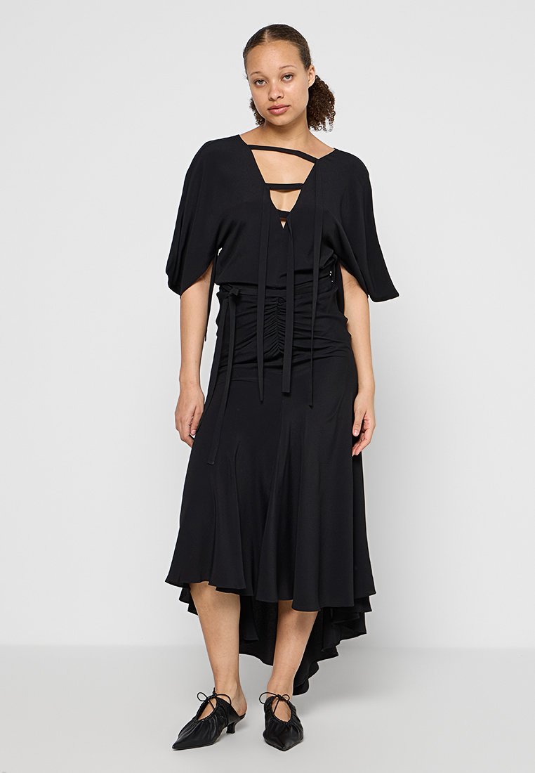 Schwarzes Kleid mit lockeren Ärmeln, V-Ausschnitt und gerafftem Bund. Mit einem ausgestellten Rock mit einem hoch-niedrig geschnittenen Saum und strukturierter Stoff. Schwarze Schnürschuhe.