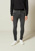Person, der einen schwarzen Kapuzenpullover, eng anliegende dunkelgraue Jeans, weiße Socken und weiße Slipper trägt, steht vor einem schlichten hellen Hintergrund.