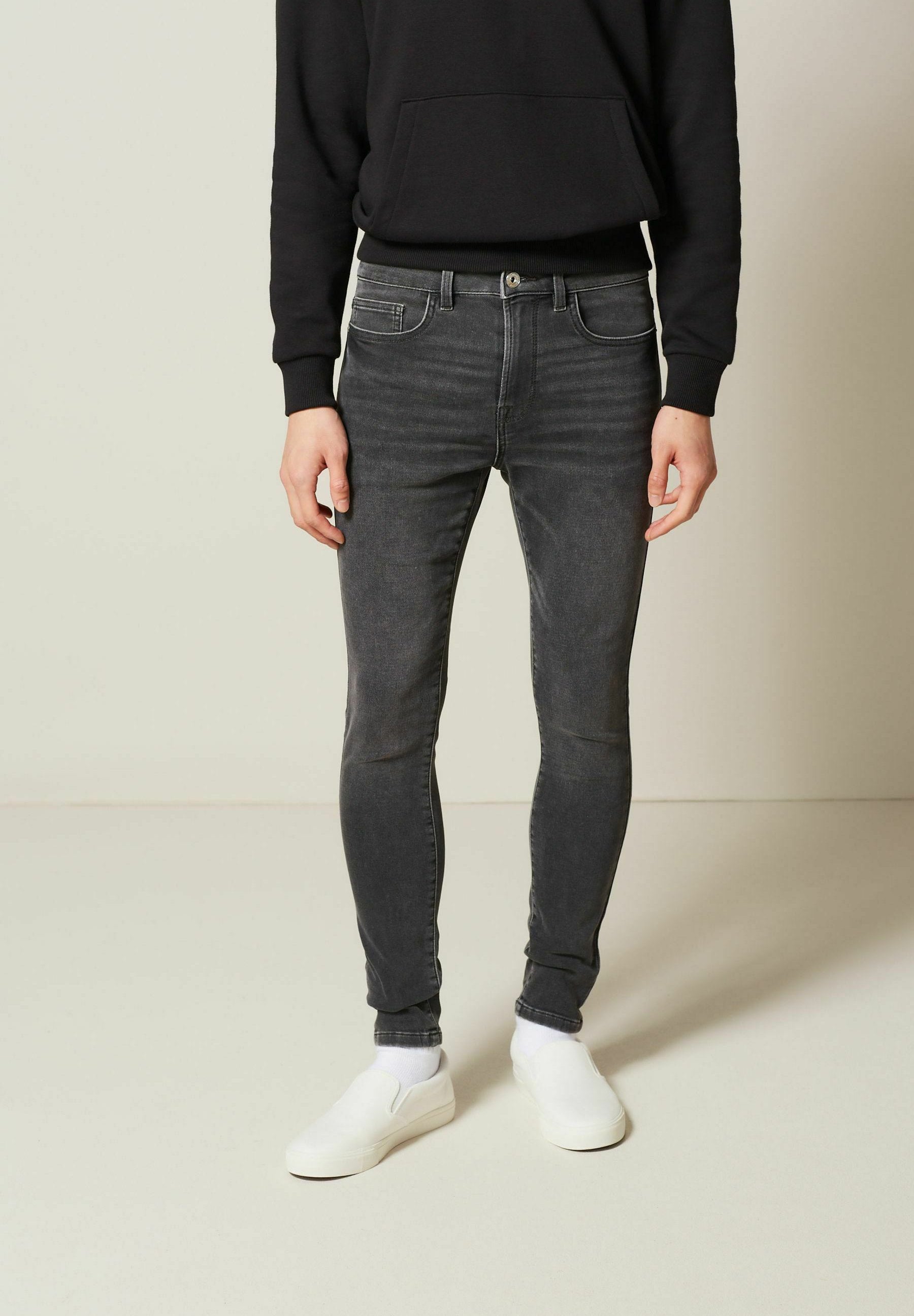 Person, der einen schwarzen Kapuzenpullover, eng anliegende dunkelgraue Jeans, weiße Socken und weiße Slipper trägt, steht vor einem schlichten hellen Hintergrund.