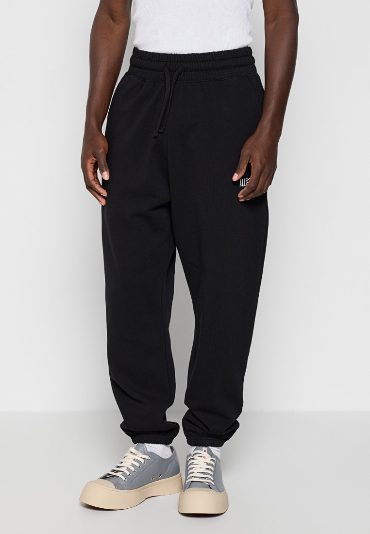 Mann trägt schwarze Sweatpants mit elastischem Bund und graue Sneakers mit weißen Schnürsenkeln, steht mit entspannten Händen neben dem Körper.