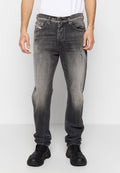 Mann trägt ausgewaschene schwarze Straight-Fit-Jeans mit leichter Abnutzung, schwarze Sneakers und ein weißes T-Shirt, steht vor einem schlichten Hintergrund.