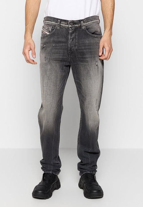 Mann trägt ausgewaschene schwarze Straight-Fit-Jeans mit leichter Abnutzung, schwarze Sneakers und ein weißes T-Shirt, steht vor einem schlichten Hintergrund.