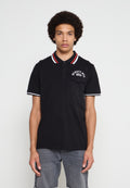 Junger Mann mit lockigem Haar, trägt ein schwarzes Poloshirt mit rot-weiß gestreiftem Kragen und graue Hose, blickt nach vorne vor einfachem Hintergrund.