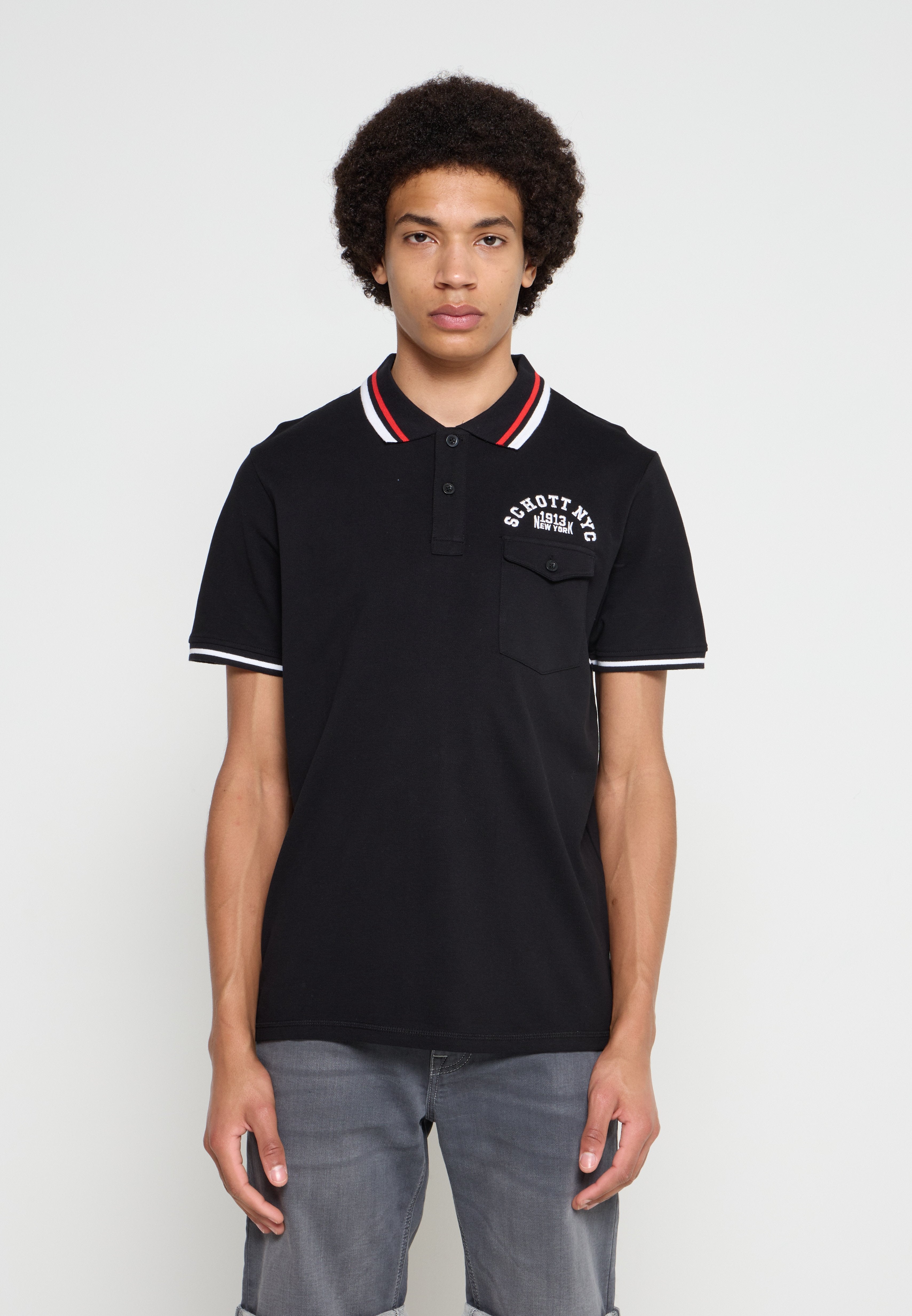 Junger Mann mit lockigem Haar, trägt ein schwarzes Poloshirt mit rot-weiß gestreiftem Kragen und graue Hose, blickt nach vorne vor einfachem Hintergrund.