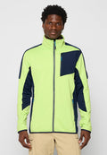 Mann in einer neon grünen und marineblauen Zip-Jacke mit Reißverschlusstaschen und beigen Cargo-Hosen, der vor einem schlichten Hintergrund steht.