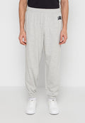 Mann trägt hellgraue, elastische Jogginghose mit Bündchen, dazu weiße Sneaker und ein schwarzes Shirt, steht vor einem schlichten Hintergrund.