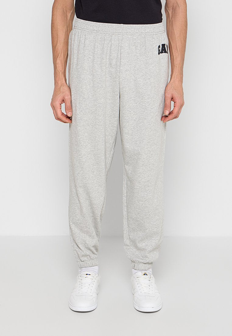 Mann trägt hellgraue, elastische Jogginghose mit Bündchen, dazu weiße Sneaker und ein schwarzes Shirt, steht vor einem schlichten Hintergrund.
