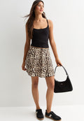 Eine Frau trägt ein schwarzes Tanktop, einen Rock mit Leopardenmuster, schwarze Sneakers und hält eine schwarze Handtasche, während sie vor einem weißen Hintergrund steht.