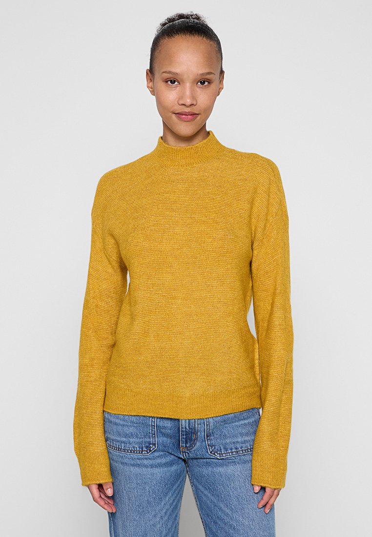 Senfgelber Strickpullover mit hohem Kragen, langen Ärmeln und legerer Passform, kombiniert mit hellblauen Jeans.