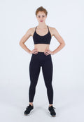 Junge Frau steht mit Händen auf den Hüften, trägt einen schwarzen Sport-BH, schwarze Leggings und schwarze Sneakers vor weißem Hintergrund.