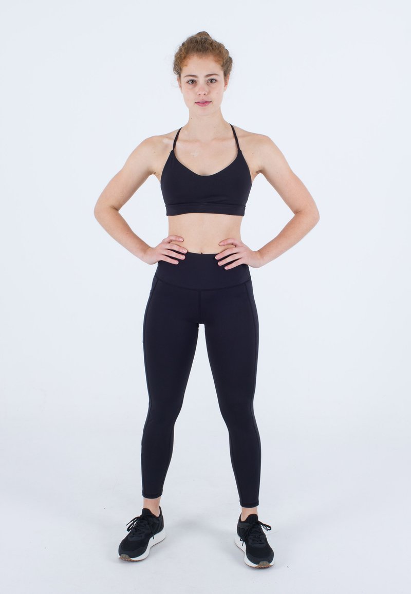 Junge Frau steht mit Händen auf den Hüften, trägt einen schwarzen Sport-BH, schwarze Leggings und schwarze Sneakers vor weißem Hintergrund.