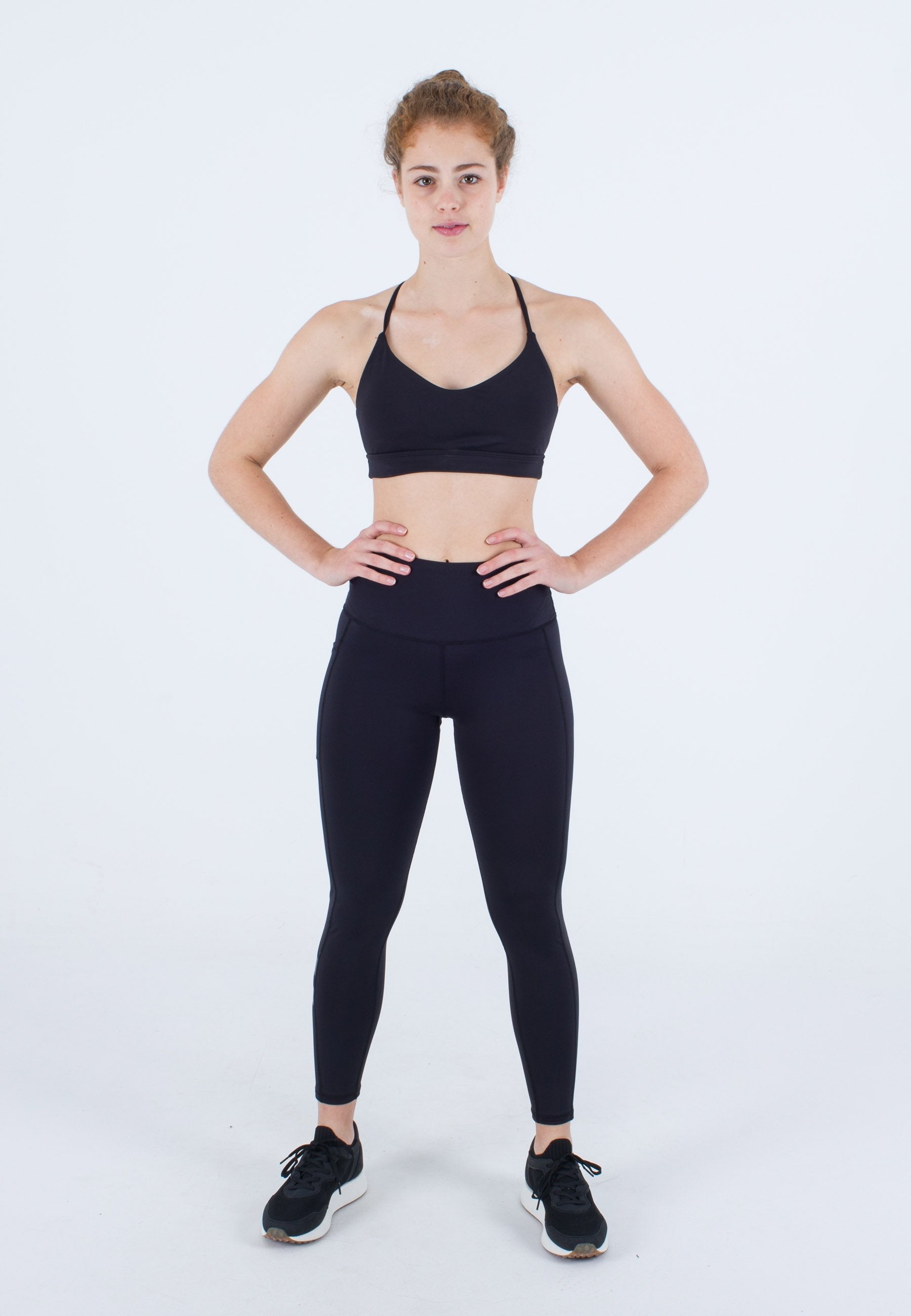 Junge Frau steht mit Händen auf den Hüften, trägt einen schwarzen Sport-BH, schwarze Leggings und schwarze Sneakers vor weißem Hintergrund.