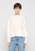 Junge Person mit kurzem braunem Haar, die einen weißen, flauschigen Pullover und hellblaue Jeans vor einem einfarbigen Hintergrund trägt.