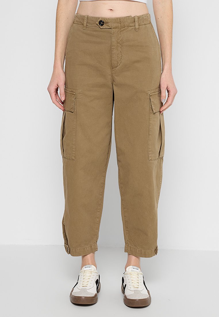 Person in braunen, taillierten Cargo-Hosen mit Klappentaschen und weißen Sneakers mit schwarzen Akzenten, stehend vor einem schlichten, weißen Hintergrund.