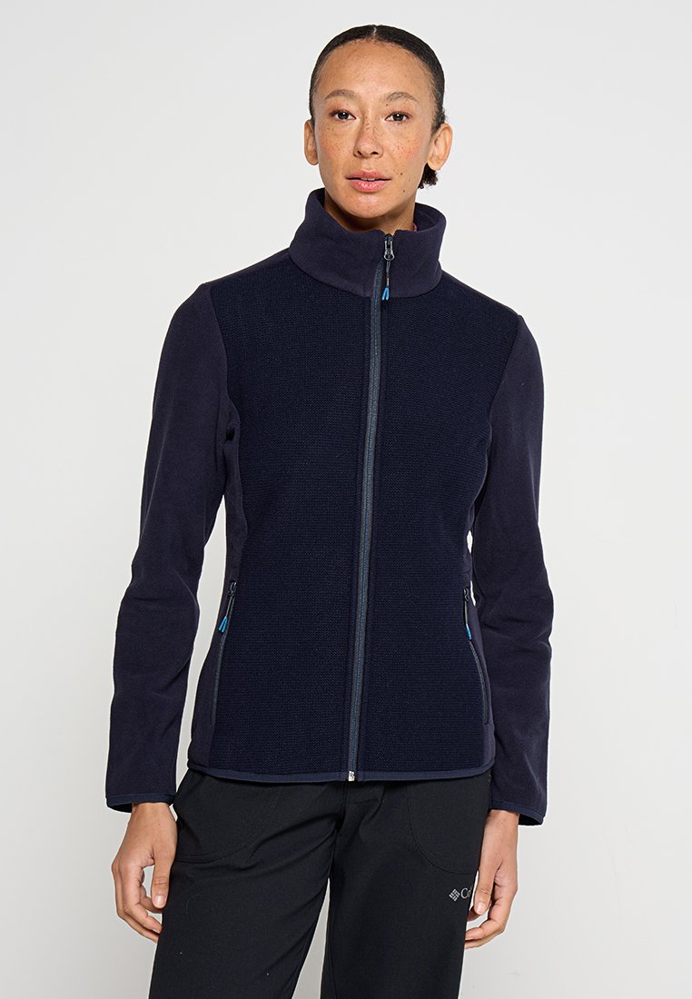Frau in einer marineblauen Fleecejacke mit Reißverschluss und hohem Kragen sowie schwarzen Hosen, steht vor einem schlichten weißen Hintergrund.