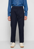 Mann trägt dunkelblaue, gerade geschnittene Hose, blaues Hemd mit Knöpfen und weiße Turnschuhe und steht vor hellem Hintergrund.