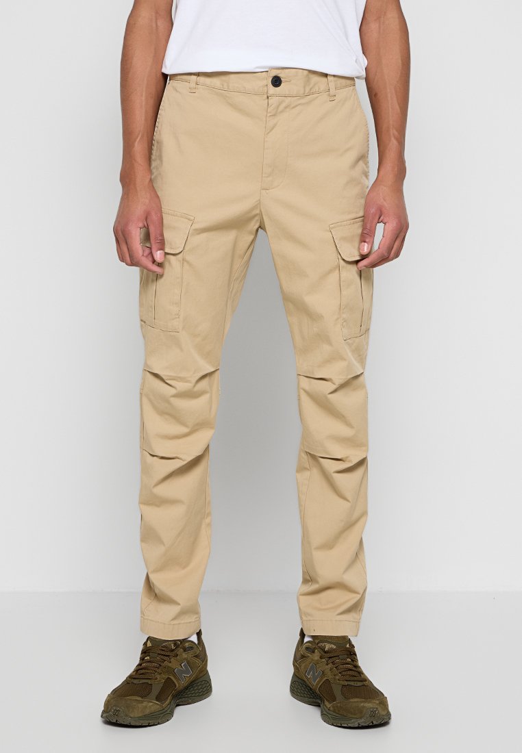Beige Cargo-Hosen aus Baumwolle, mit zwei Seitentaschen, einer lockeren Passform und einem geraden Bein-Design. Kombiniert mit grünen Sportschuhen.